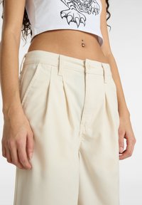 Pantaloni plissettati di colore crema con chiusura a bottone e passanti per cintura. Tessuto morbido e vestibilità rilassata, dotati di tasche laterali.