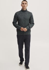 Dunkelgrüner, kabelgestrickter Rollkragenpullover mit strukturiertem Muster, kombiniert mit dunkelblauen Hosen und schwarzen Sneakers mit weißen Sohlen.
