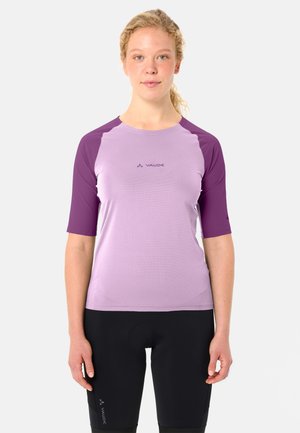 Jonge vrouw met krullend blond haar, gekleed in een tweekleurig paars wielertricot en zwarte wielerbroek, staand tegen een witte achtergrond.