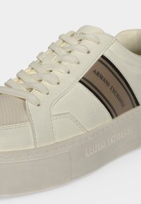 Baskets blanches en cuir avec détail en mesh beige, lacets beiges et panneau en tissu rayé noir avec logo Armani Exchange sur le côté et la semelle.