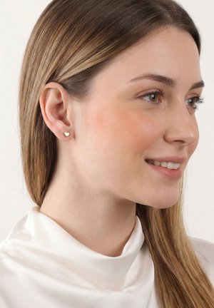 Jeune femme aux longs cheveux raides portant de petites boucles d'oreilles en forme de cœur en or et un haut blanc à col montant, souriant légèrement sur le côté.