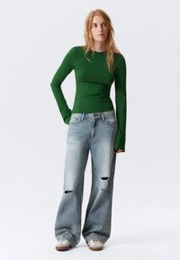 Grünes Langarmshirt, kombiniert mit hellblauen, weit geschnittenen Jeans mit zerstörten Details. Das Outfit wird mit beigen Sneakers abgerundet.