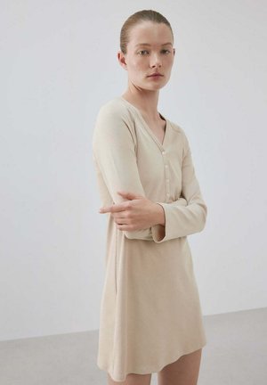 Jeune femme à la peau claire et aux cheveux tirés en arrière porte une robe beige à manches longues et boutonnée, debout devant un fond gris clair uni.