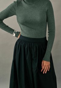 Pull à col roulé côtelé vert associé à une jupe plissée noire. La tenue présente des manches ajustées et un tissu texturé.