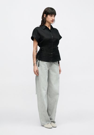 Μαύρο πουκάμισο με κουμπιά, φτιαγμένο από υφή υφάσματος, με κοντά μανίκια και λεπτομέρειες με κορδόνια, συνδυασμένο με ανοιχτό γκρι παντελόνι με φαρδιά πόδια.