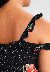 Czarna sukienka w kropki z falbaną na ramiączku, kwiatowymi akcentami w kolorze czerwonym i zielonym oraz suwakiem z tyłu.