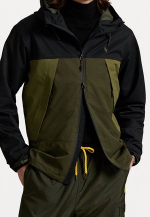 Mand iført en sort og olivengrøn to-farvet jakke med hætte, kombineret med olivengrønne bukser med en gul snor til justering.