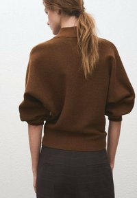 Frau mit hellbraunem Pferdeschwanz, die einen braunen Strickpullover mit Puffärmeln und dunkle karierte Hose trägt, steht vor einer schlichten weißen Wand.