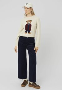 Pull en maille crème avec un motif d'ours, associé à un pantalon large en velours côtelé bleu marine et des chaussures à lacets marron avec une semelle épaisse.