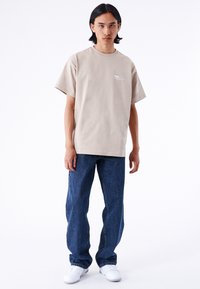 Camiseta de algodón beige con mangas cortas, ajuste holgado y un sutil diseño de texto en el pecho, combinada con jeans de mezclilla azules y zapatillas blancas.