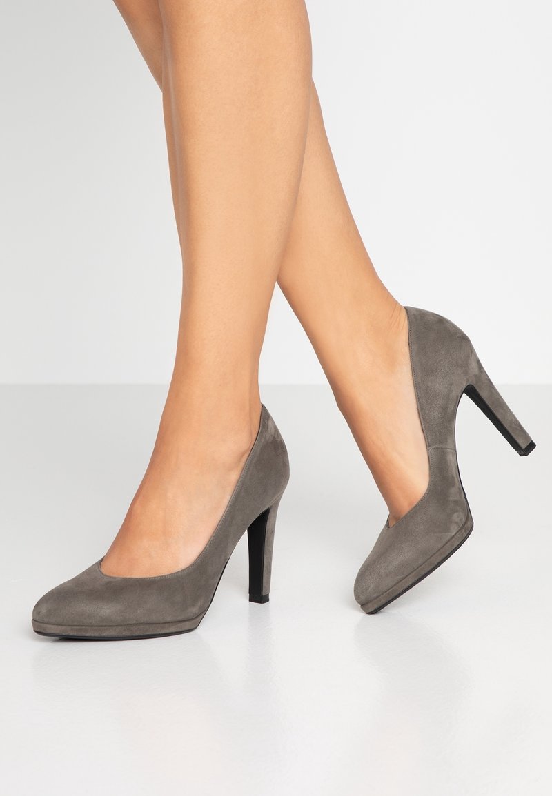 Jambes croisées portant des escarpins en daim gris à bouts ronds et talons aiguilles noirs sur un sol blanc.