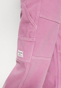 Närbild av rosa denimbyxa ficka med vit BDG Jeans-varumärkesetikett och detaljerad vit sömnad.