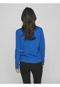 Blå stickad tröja med strukturerat mönster, lång ärm och ribbade muddar och nedkant, tillsammans med svarta byxor, sedd bakifrån.