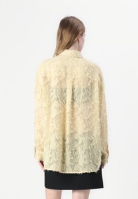 Camicia gialla, trasparente, con texture frangiata e un fit rilassato. Presenta maniche lunghe e un colletto, scendendo appena sotto la vita.