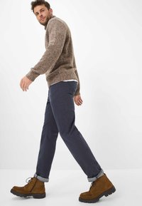 Maglione marrone con texture, pantaloni blu navy e stivali marroni con lacci e suola spessa. I polsini arrotolati dei pantaloni mostrano un motivo a scacchi.