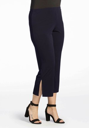 Pantalons cropped navy avec fentes latérales, fabriqués en tissu extensible. Associés à des sandales à talons noires, mettant en valeur une texture lisse et une coupe ajustée.