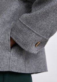 Casaco de tecido cinza com uma textura suave e escovada; apresenta um punho dobrado com um acessório de botão metálico redondo e abertura de bolso lateral.