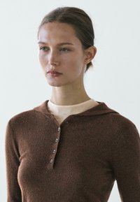 Woman with light skin and light brown hair, wearing a brown ribbed button-up sweater over a beige top, looking slightly to the side against a plain background.