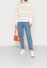 Weißer Strickpullover mit orangefarbenen, blauen und gelben Wellenmustern; blaue Jeans; orangefarbige Handtasche; und weiße Sneakers.