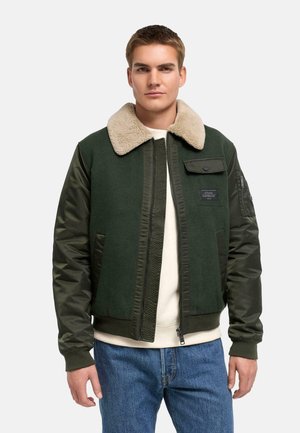 Jeune homme portant un bomber vert foncé avec un col en peau retournée beige, chemise blanche en dessous et jean bleu, debout devant un fond uni.