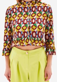 Multifarvet cropped bluse med geometriske mønstre i rød, grøn, gul og blå, med knaplukninger og flagrende manchetter.