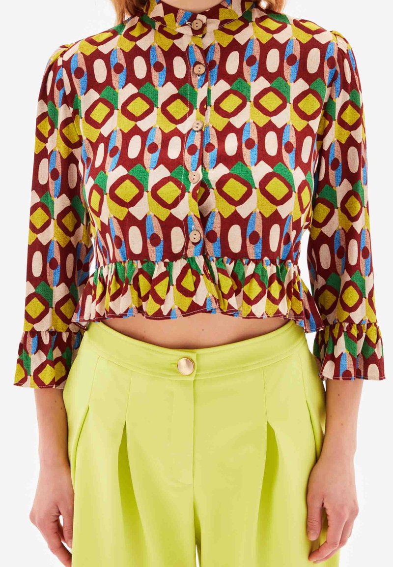 Multifarvet cropped bluse med geometriske mønstre i rød, grøn, gul og blå, med knaplukninger og flagrende manchetter.