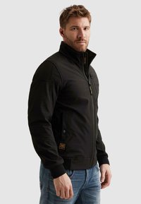 Uomo che indossa una giacca nera con zip e colletto alto e jeans blu, in piedi contro uno sfondo chiaro e uniforme.