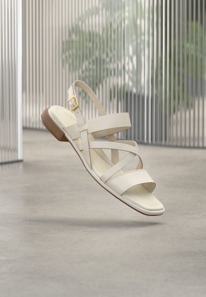 Sandale plate en cuir beige avec des brides croisées, boucle dorée et talon bas en bois flottant au-dessus d'un sol en béton avec un arrière-plan flou de barres métalliques verticales.