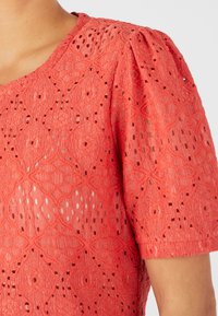 Blusa in pizzo corallo con maniche corte a palloncino. Presenta un motivo a diamante e floreale con una finitura testurizzata e piccoli tagli decorativi.