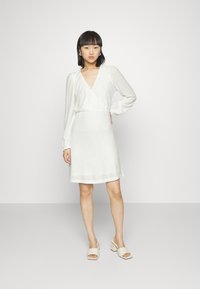 Vestido branco texturizado com decote em V, mangas longas e cintura elástica. Usado com sandálias de salto bloco em bege.