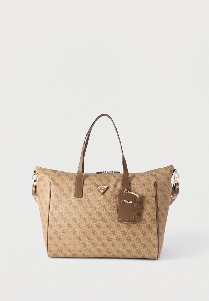 Καφέ τσάντα ώμου με σχέδιο, διπλά χερούλια, μπροστινό τριγωνικό λογότυπο και προσαρτημένη ορθογώνια ετικέτα που γράφει "GUESS" σε απλό φόντο.