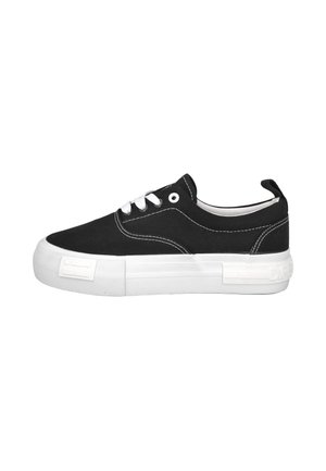 Zapatilla deportiva negra de caña baja con cordones blancos, suela blanca gruesa, costuras visibles y una pestaña para tirar en el talón sobre un fondo blanco liso.