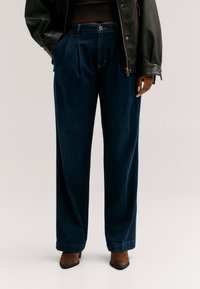 Personne portant un jean bleu foncé à jambes larges, des bottes marron à bout pointu, un top noir court et une veste en cuir noire avec les mains partiellement dans les poches.