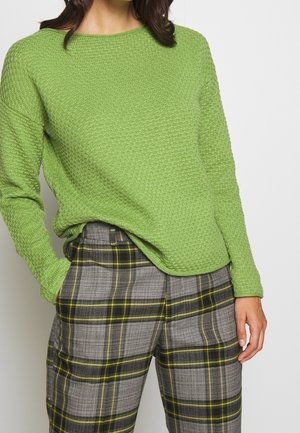 Femme portant un pull vert clair texturé et un pantalon à carreaux gris avec des lignes jaunes et noires, main dans la poche sur un fond uni.