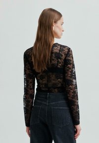 Kobieta z długimi brązowymi włosami, ubrana w czarną koronkową bluzkę z długim rękawem i ciemne, wysokiej talii dżinsy, stoi bokiem na neutralnym tle.