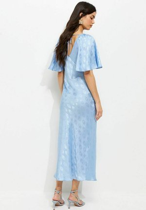 Frau mit langen dunklen Haaren, die ein hellblaues Midikleid mit flatternden Ärmeln, offenem Rücken mit Bindeband und silbernen Riemchenabsätzen trägt, von hinten gesehen.