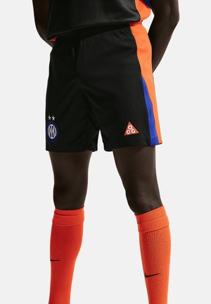 Mand iført sorte sportsshorts med blå og orange striber og orange knælange sokker med sorte Nike-swooshes.