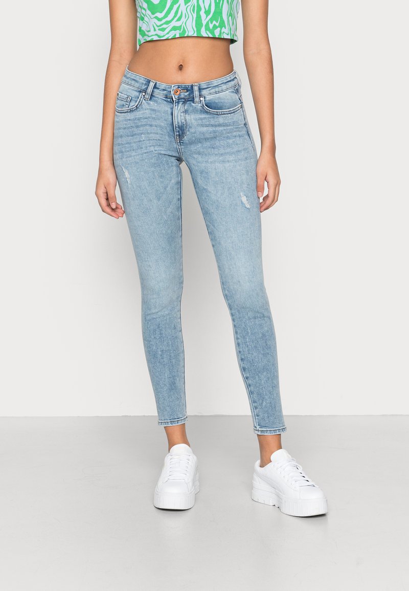 Jeans skinny em azul claro com detalhes desgastados, design de cinco bolsos, bainhas desfiadas e cintura média, combinados com ténis de plataforma brancos.