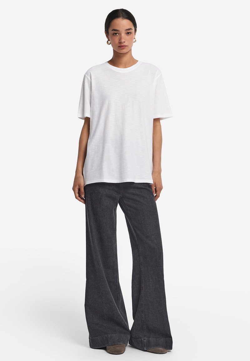 Hvid, kortærmet t-shirt lavet af blødt bomuld, parret med højtaljede, vide, mørk denim jeans og grå fodtøj. Enkel design, ingen mønstre.