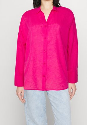Femme portant une chemise à manches longues rose vif boutonnée, avec un jean bleu clair, debout devant un fond clair uni.