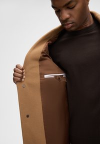 Tanskleurige wolletjes met een gladde bruine voering, voorzien van een borstzak met een wit en bruin accent. De jas is iets open, waardoor de binnenkant zichtbaar is.