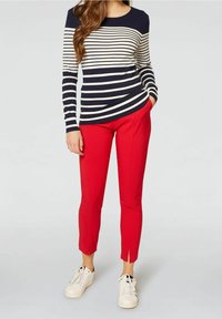 Un pull rayé bleu marine et crème assorti à un pantalon rouge slim avec des fentes sur les côtés, porté avec des baskets blanches sur un fond clair.