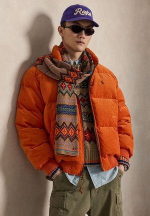 Mann trägt orangefarbene Steppjacke, gemusterten Schal, Strickpullover, lila Mütze, Sonnenbrille, blaues Hemd und grüne Cargohosen mit den Händen in den Taschen vor einer beigen Wand.