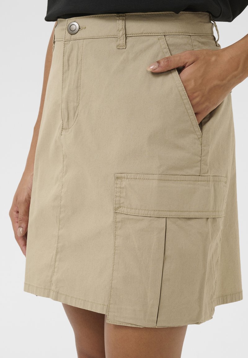 Beige bavlněná sukně se bočními kapsami, s jedním předním záhybem a kovovým knoflíkem; hladká textura a délka ke kolenům.