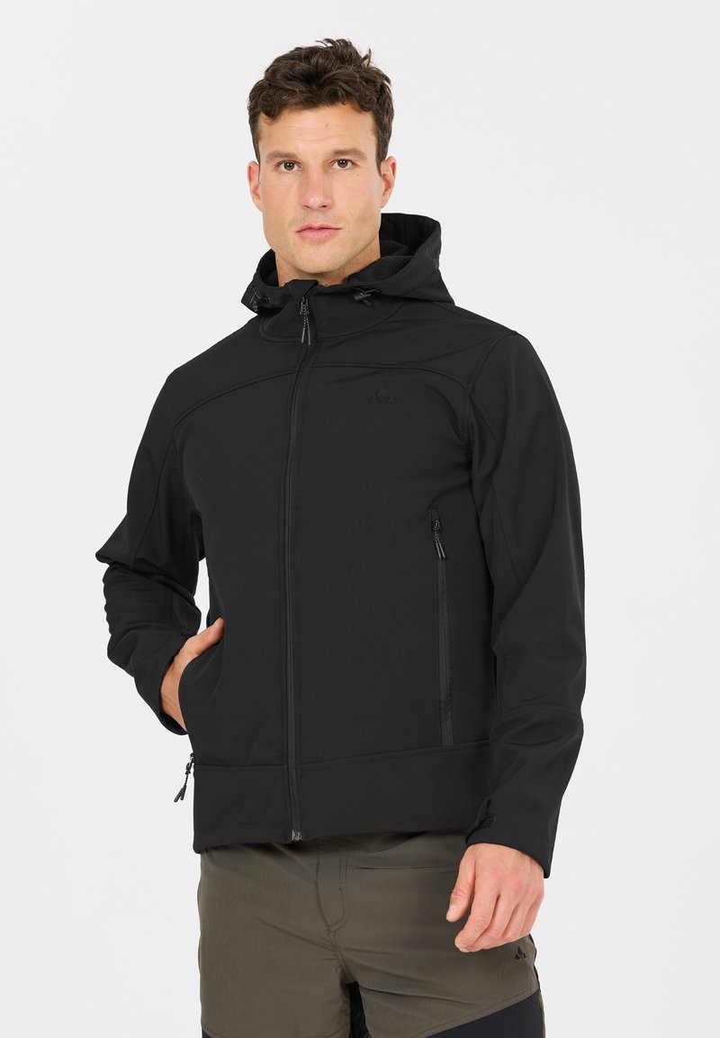 Homme portant une veste softshell noire à capuche avec des poches zippées et un pantalon vert olive, debout devant un fond uni.