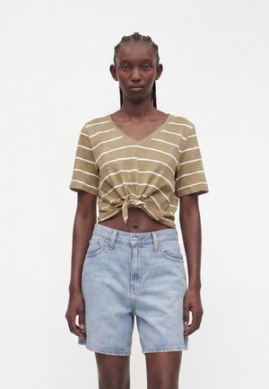 Femme aux cheveux tressés porte un crop top à manches courtes beige et blanc rayé noué à la taille et un short en jean taille haute bleu clair.