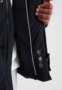 Chaqueta de plumas negra con cierre de cremallera y botón, con ribetes blancos, una textura acolchada y símbolos de cuidado que indican propiedades a prueba de viento y repelente al agua.