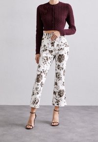 Burgunderfarbener gestrickter kurzer Cardigan mit Vorderknöpfen und weißer Hose mit floralem Muster in Brauntönen, kombiniert mit schwarzen, hochhackigen Riemchenschuhen.