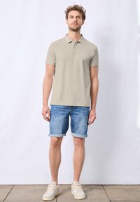 Světlé béžové polo tričko se třemi knoflíky, spárované s džíny šortkami s ohrnutými rukávy. Nosené se světlými teniskami na texturované podlaze.