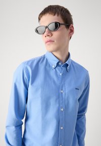 Lichtblauw button-up shirt met lange mouwen en kraag, voorzien van een klein zwart logo. Zwarte ovale zonnebril met dunne rand.